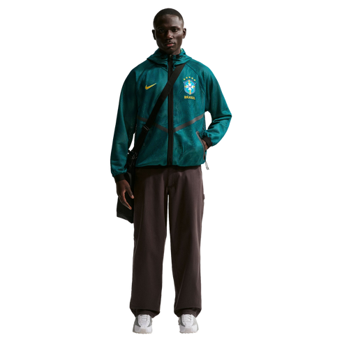 A young Black man stands against a black background wearing a teal mesh Nike zip-up hoodie featuring the Brazil national football team crest, dark brown wide-leg trousers, and white sneakers. He has a black messenger bag slung across his chest.