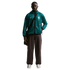 A young Black man stands against a black background wearing a teal mesh Nike zip-up hoodie featuring the Brazil national football team crest, dark brown wide-leg trousers, and white sneakers. He has a black messenger bag slung across his chest.
