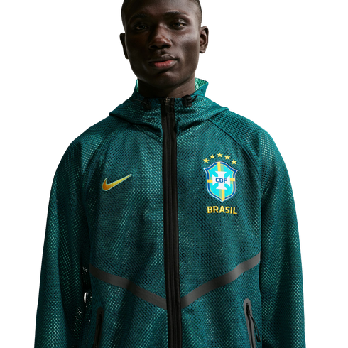 A waist-up portrait of a young Black man wearing a dark teal mesh Nike zip-up hoodie. The hoodie features a yellow Nike swoosh on one side and the Brazil national football team crest with "BRASIL" printed below it on the other. He is looking directly at t