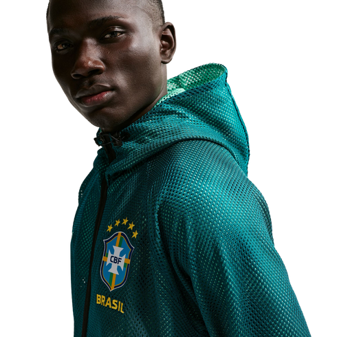 A close-up shot of a man with dark skin looking towards the camera against a solid black background. He is wearing a teal, mesh, zip-up hoodie with the Brazilian football confederation (CBF) crest on the chest, featuring five yellow stars above it and the