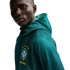 A close-up shot of a man with dark skin looking towards the camera against a solid black background. He is wearing a teal, mesh, zip-up hoodie with the Brazilian football confederation (CBF) crest on the chest, featuring five yellow stars above it and the