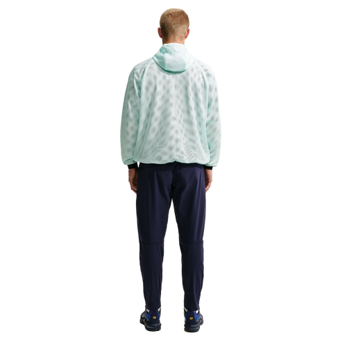 A man stands with his back to the camera, wearing a light mint green mesh hoodie, navy blue pants, and blue sneakers against a black background.
