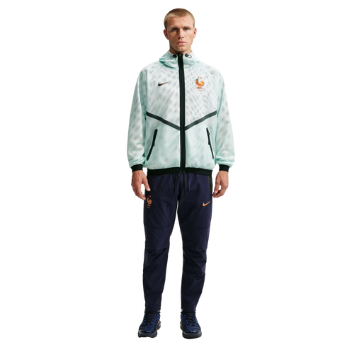 A man stands against a solid black background wearing a Nike tracksuit for the French national football team. He is wearing a light mint green, mesh-textured hooded jacket with black accents and navy blue track pants with orange logos.