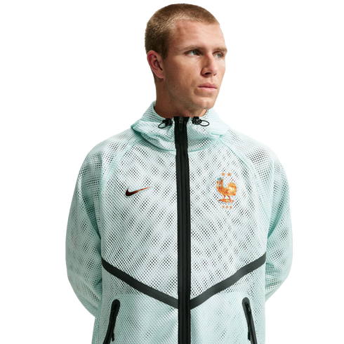 A man wearing a light mint green, mesh Nike zip-up hoodie with the French Football Federation (FFF) crest and black chevron detailing, set against a solid black background.
