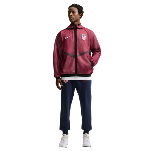 A full-length photo of a man modeling Nike USA soccer apparel against a black background. He wears a maroon hooded jacket with a white swoosh and USA crest, navy joggers with matching logos, and white sneakers.