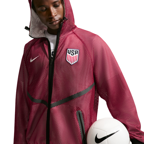 A young Black man looking down while wearing a maroon mesh hooded jacket featuring a "USA" soccer crest and a white Nike swoosh. He holds a white Nike soccer ball against his hip, set against a solid black background.