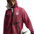 A young Black man looking down while wearing a maroon mesh hooded jacket featuring a "USA" soccer crest and a white Nike swoosh. He holds a white Nike soccer ball against his hip, set against a solid black background.