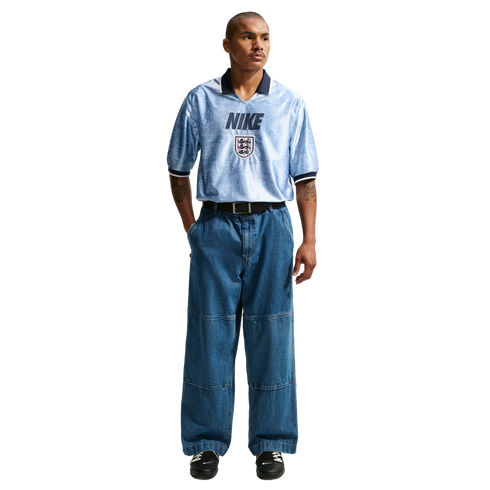 A man models a light blue Nike England jersey tucked into baggy wide-leg blue jeans and black sneakers against a plain white background.