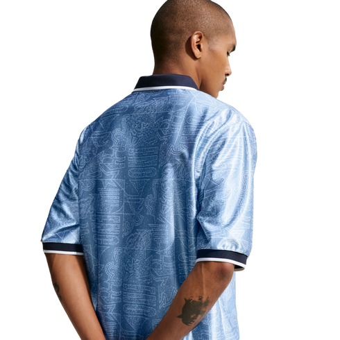 Back view of a man in a patterned light blue polo shirt with navy and white trim, standing against a white background with his hands behind his back.