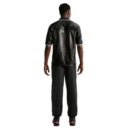 Rear view of a man in a black polo shirt with a Statue of Liberty graphic, black pants, and dark sneakers against a white background.