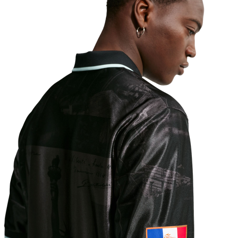 Profile of a Black man from behind, wearing a shiny black polo shirt with historical graphic prints and a small flag patch on the sleeve.