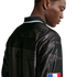 Profile of a Black man from behind, wearing a shiny black polo shirt with historical graphic prints and a small flag patch on the sleeve.
