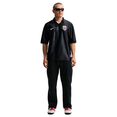 A man modeling a black Nike Norway football jersey with a Nordic knot pattern, black sweatpants, sunglasses, and red and white sneakers against a white background.