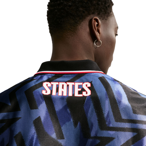 Rear view of a person with short dark hair wearing a blue patterned jersey with "STATES" in white and red text on the back. They are wearing a silver hoop earring.