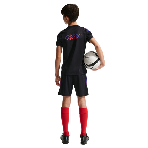 Back view of a young boy in a black soccer kit and red socks, holding a white and black soccer ball against a solid black background.