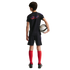 Back view of a young boy in a black soccer kit and red socks, holding a white and black soccer ball against a solid black background.
