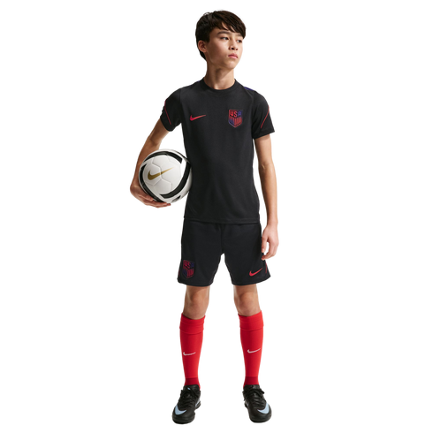 A young boy in a black USA soccer kit and red socks holds a soccer ball against a solid black background.