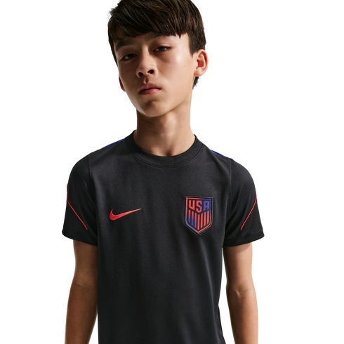 A young boy with short brown hair wearing a black Nike USA soccer jersey, looking at the camera against a solid black background.