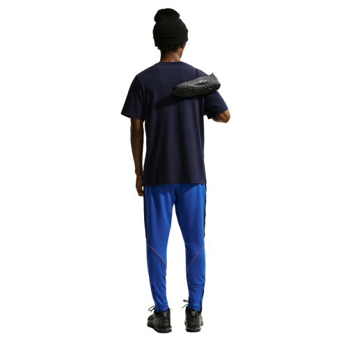Rear view of a person wearing a navy t-shirt, bright blue track pants with orange trim, and a black beanie. They have a black Nike soccer cleat slung over their shoulder against a solid black background.