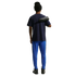 Rear view of a person wearing a navy t-shirt, bright blue track pants with orange trim, and a black beanie. They have a black Nike soccer cleat slung over their shoulder against a solid black background.