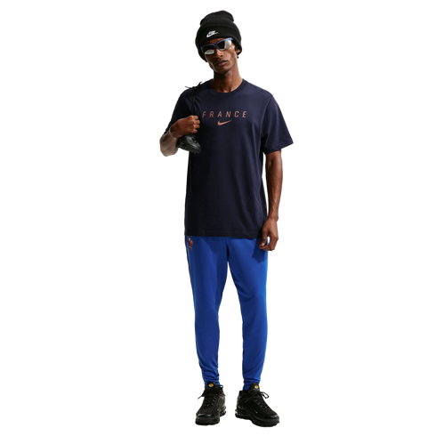A full-body photo of a man standing against a solid black background. He is wearing a dark navy blue Nike t-shirt with "FRANCE" printed on the chest, bright blue athletic pants, black sneakers, a black Nike beanie, and blue-tinted sunglasses. He is holdin