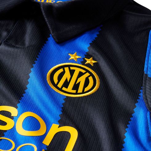 Close-up of an Inter Milan jersey with blue and black jagged stripes. It features the team's gold crest under two stars and part of a yellow sponsor logo.