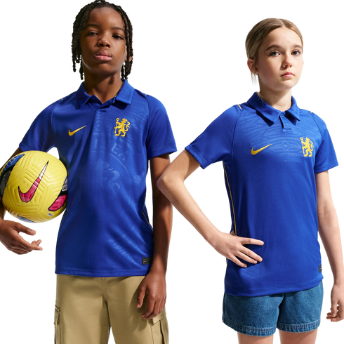 Two children wearing blue Nike Chelsea FC polo jerseys pose against a white background. The boy on the left holds a yellow soccer ball.