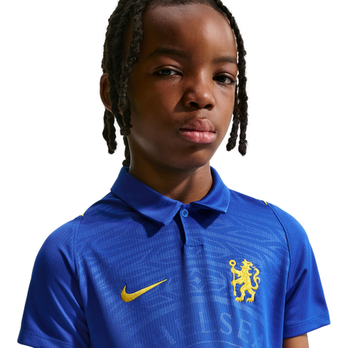 Portrait of a young person with braided hair wearing a blue Chelsea FC polo shirt with a yellow lion crest, against a white background.