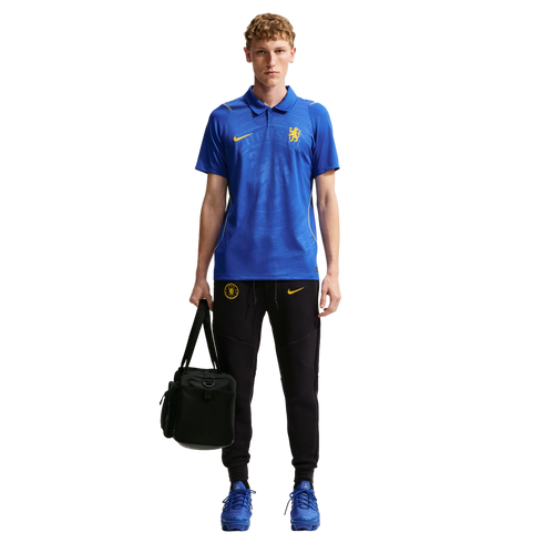 A person wearing a blue Chelsea FC polo shirt, black Nike joggers, and blue sneakers, holding a black gym bag.