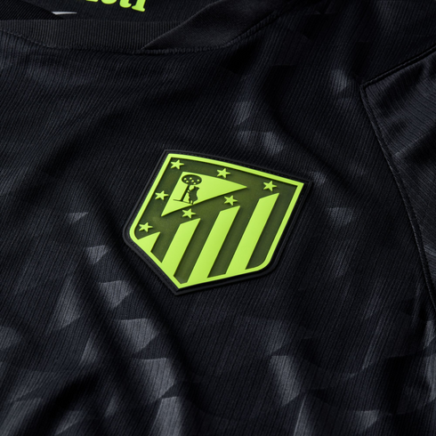Close-up of a black soccer jersey with a lime green Atlético Madrid crest. The fabric features a subtle tonal geometric pattern.