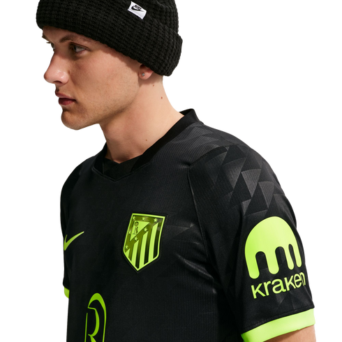 A man in profile wearing a black Nike beanie and a black Atletico Madrid jersey with neon green logos and a Kraken sponsor patch on the sleeve.
