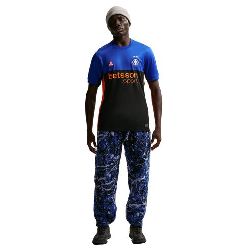 A man models a blue and black Inter Milan x Nike ACG jersey, blue patterned trousers, a grey beanie, and dark sneakers against a black background.