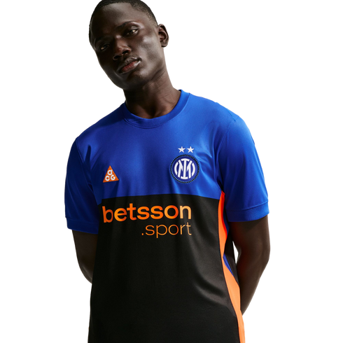 A man in a blue and black Inter Milan training jersey with orange "betsson.sport" and ACG logos. He poses with hands behind his back against a black background.