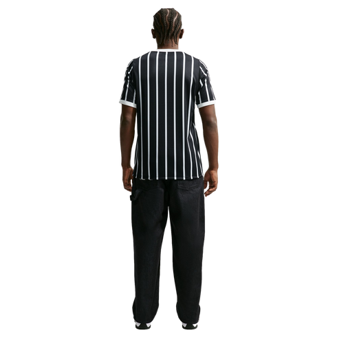 Rear view of a man with braided hair wearing a black and white vertical striped T-shirt and black trousers against a solid black background.