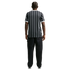 Rear view of a man with braided hair wearing a black and white vertical striped T-shirt and black trousers against a solid black background.