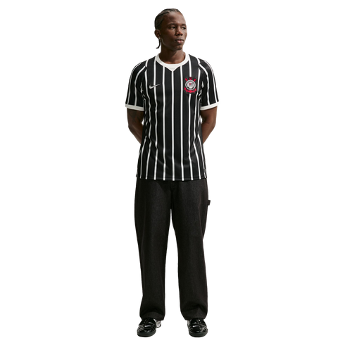 Full-length shot of a man in a black and white striped Corinthians soccer jersey, black pants, and black sneakers, standing against a solid black background.