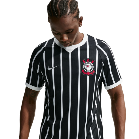 A person with braided hair looks down, wearing a black and white vertically striped Nike soccer jersey with a red and white crest. The background is solid black.