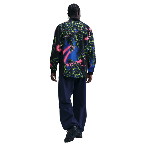 Rear view of a person with cornrows walking away, wearing a neon-patterned black shirt, navy trousers, and chunky black sneakers against a black background.