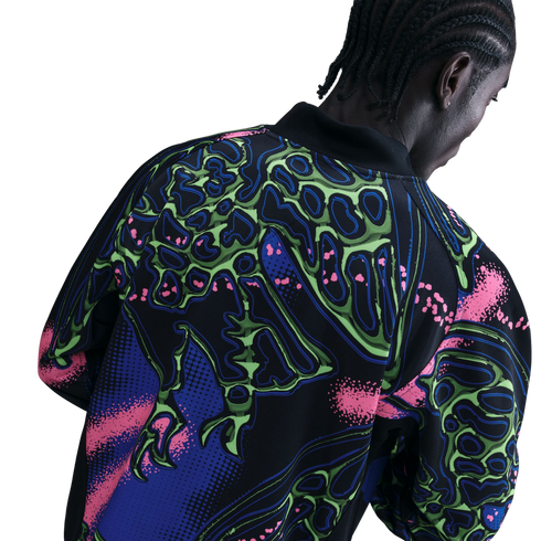 Rear view of a person with braided hair wearing a black jacket with a vibrant neon green, pink, and blue abstract skeletal pattern.