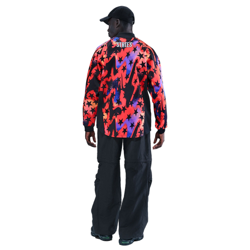 Rear view of a man wearing a black cap, a multi-colored star-patterned shirt with "STATES" on the back, and baggy black pants against a black background.