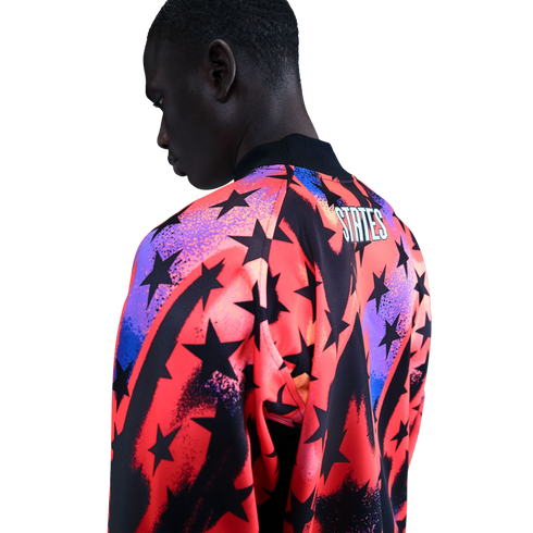 A man from the back wearing a vibrant jacket with black stars over red, orange, and purple gradients. The word "STATES" is printed on the upper back. Black background.