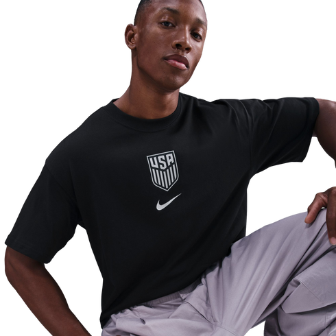 A man wearing a black Nike USA soccer T-shirt and grey cargo pants poses against a black background.