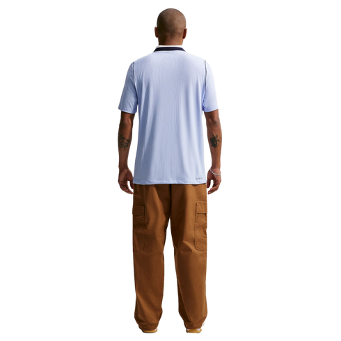 A man standing with his back to the camera against a solid black background, wearing a light blue ribbed short-sleeved shirt and baggy brown cargo pants. He has tattoos on both of his forearms.