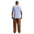 A man standing with his back to the camera against a solid black background, wearing a light blue ribbed short-sleeved shirt and baggy brown cargo pants. He has tattoos on both of his forearms.