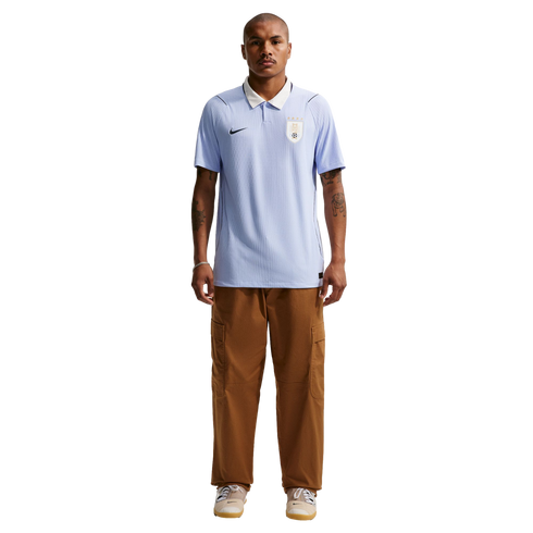 Full-body portrait of a man wearing a light blue Uruguay national team soccer jersey with a white collar, paired with brown cargo pants and beige sneakers against a solid black background.
