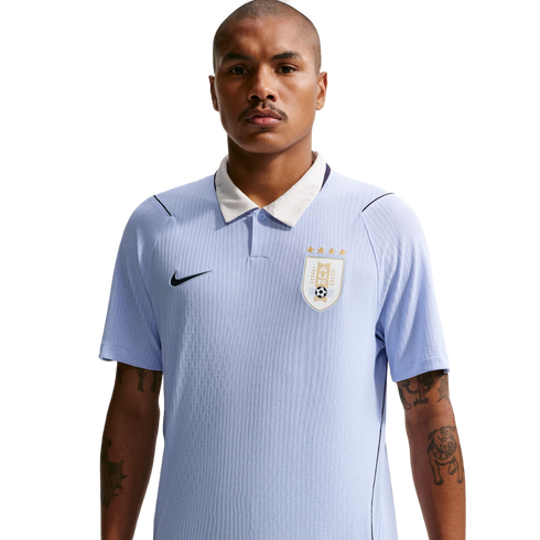 A man with a mustache and tattooed forearms wears a light blue Uruguay national team soccer jersey with a white collar against a solid black background.