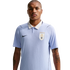 A man with a mustache and tattooed forearms wears a light blue Uruguay national team soccer jersey with a white collar against a solid black background.