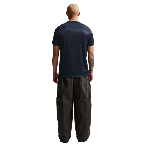 A rear view of a bald man wearing a navy blue t-shirt and oversized dark cargo pants, standing against a solid black background.