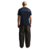 A rear view of a bald man wearing a navy blue t-shirt and oversized dark cargo pants, standing against a solid black background.