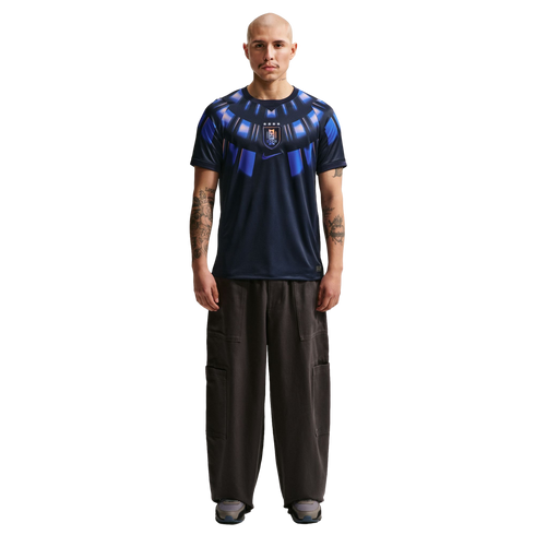 A full-body studio portrait of a bald man with arm tattoos standing against a solid black background. He is wearing a dark navy blue Nike t-shirt featuring a bright blue radial graphic around the collar and a center-chest Uruguay national team crest. His
